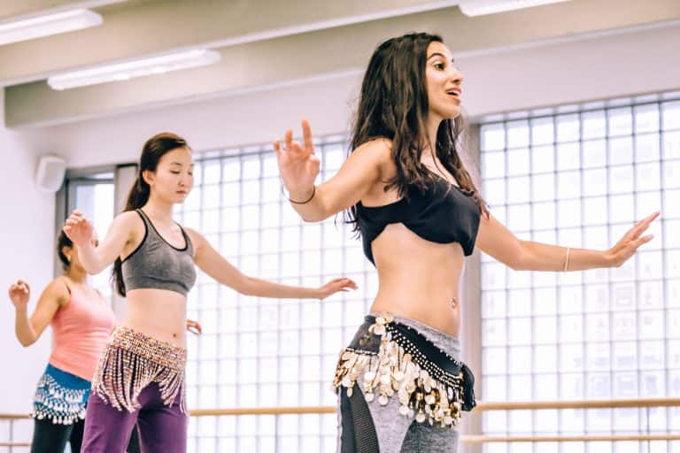 A woman teaching belly dance moves to a group of students in a studio. Belly dancing is a fun and healthy activity that improves posture, flexibility, and coordination.